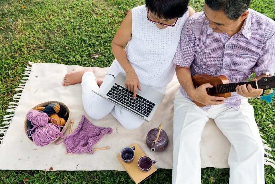 an older couple outside, sitting on a blanket, with a computer, ukelele, tea cups, and a bowl of yarn