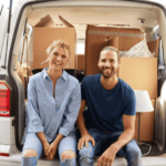 A couple sitting on the end of a filled moving van with an open trunk.