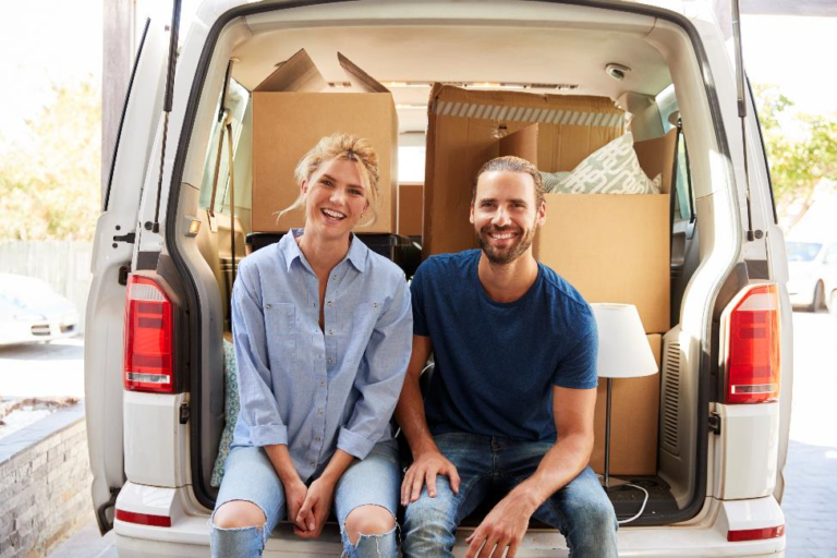 A couple sitting on the end of a filled moving van with an open trunk.