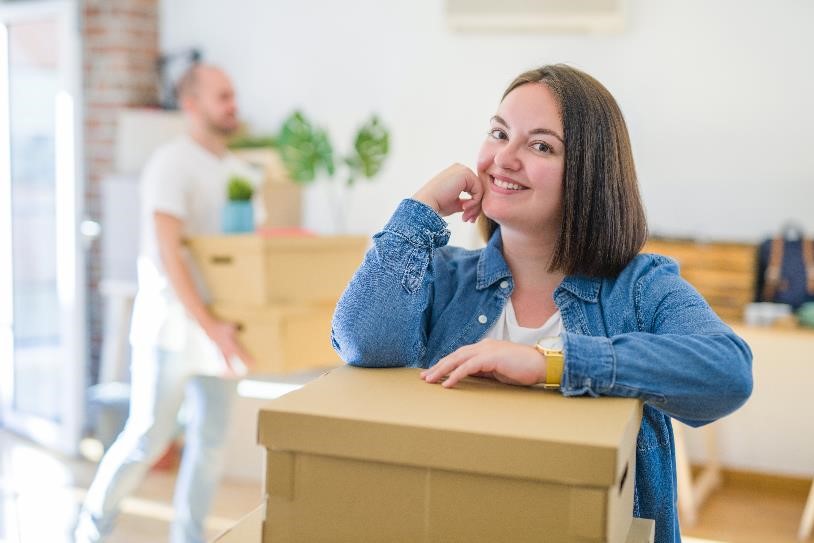A person leaning over a moving box in a home while someone carries a moving box in the background.