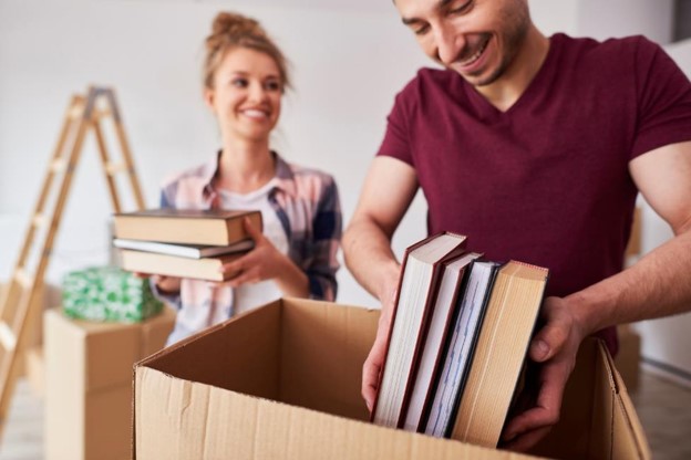 A person placing books into a moving box while someone else brings additional books to them.