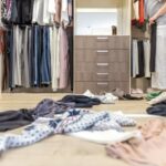 A person standing in an open closet space, cluttered with garments on the floor.