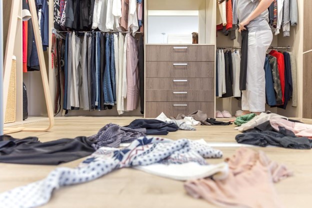 A person standing in an open closet space, cluttered with garments on the floor.