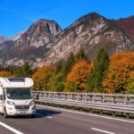 A RV drives along the highway surrounded by mountains and trees with changing leaves from green to orange.