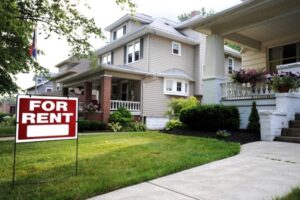 A grey home with a red "For Rent" sign posted in the front yard