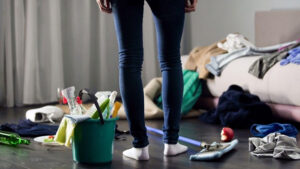 A person stands in a cluttered room next to a bucket of cleaning supplies and a mop