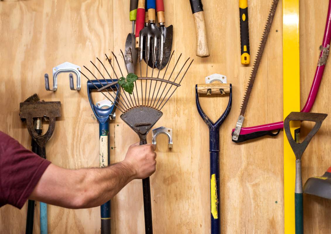 Man's arm removing metal rake from a wooden wall with various garden tools hanging organized.