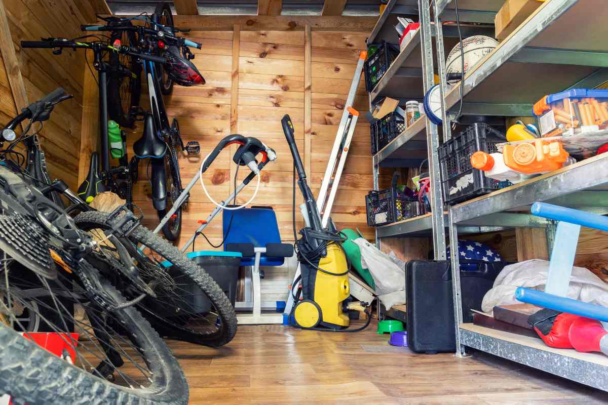 Unorganized storage in a suburban home featuring a variety of items on shelves and the floor, including bikes, exercise equipment, a ladder, and gardening tools.