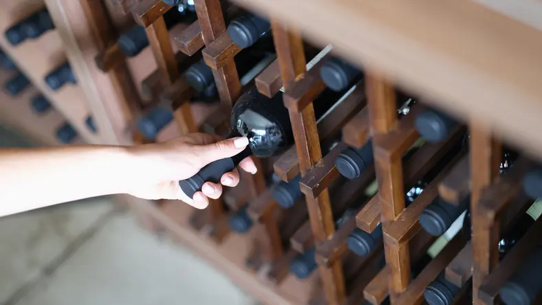 A woman’s hand selecting a wine bottle from a wooden wine rack.