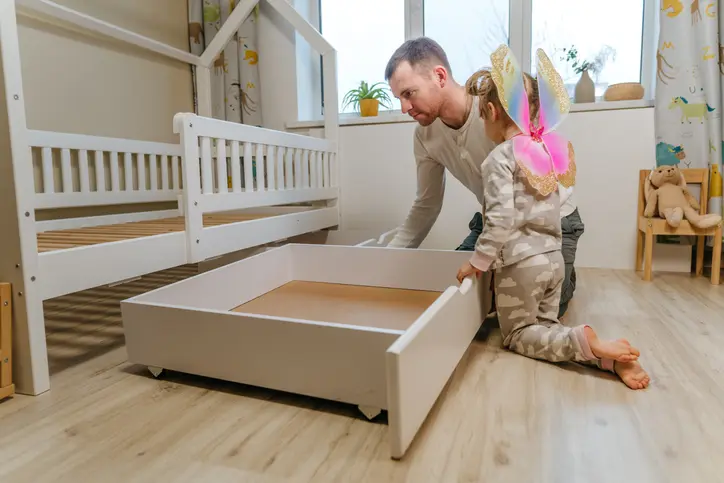 A man and young child assembling a white pullout storage drawer under a bed in a bright, organized children's bedroom.