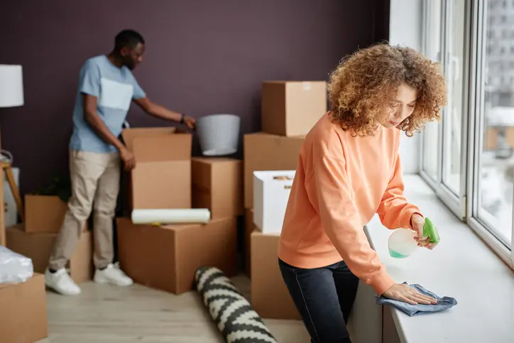 A woman cleans a window ledge with a spray bottle while a man packs boxes.