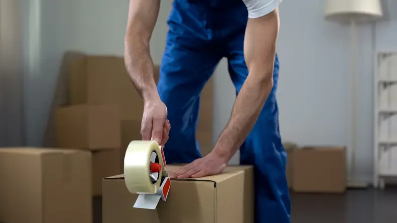 A person in blue overalls is sealing a cardboard moving box with packing tape in a room filled with boxes.