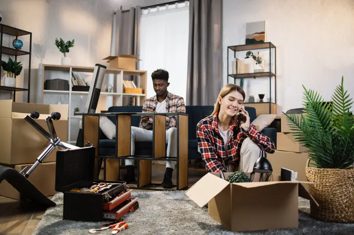 A couple packing and organizing in a living room filled with boxes, tools, and furniture during their apartment move.