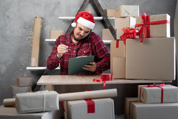 A man in a Santa hat and flannel shirt checks a clipboard with cardboard boxes tied with red ribbon surrounding him.