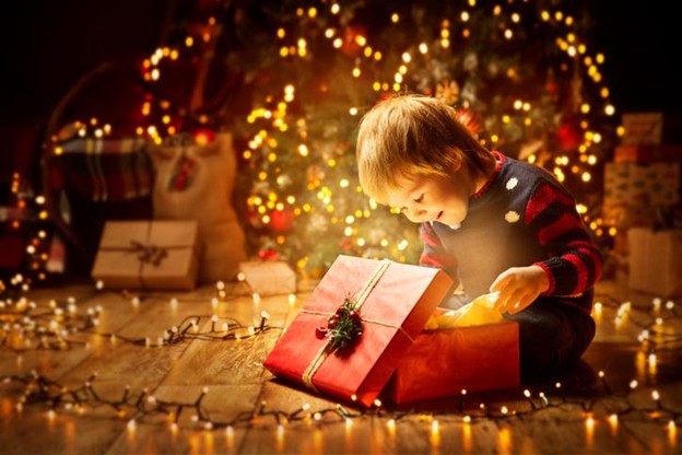 A smiling young blonde child opens a red gift with twinkling lights surrounding him and a Christmas tree behind.