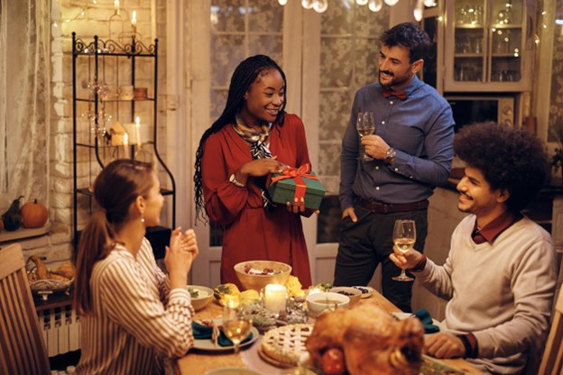 A group of four friends gather around the table for a holiday meal with a young woman holding a gift box.