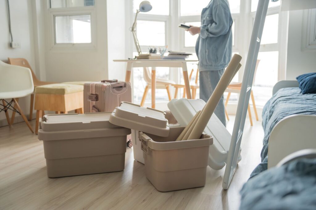 A woman in denim packs items into bins and a suitcase for a move, with chairs, a bed, and a table in the room